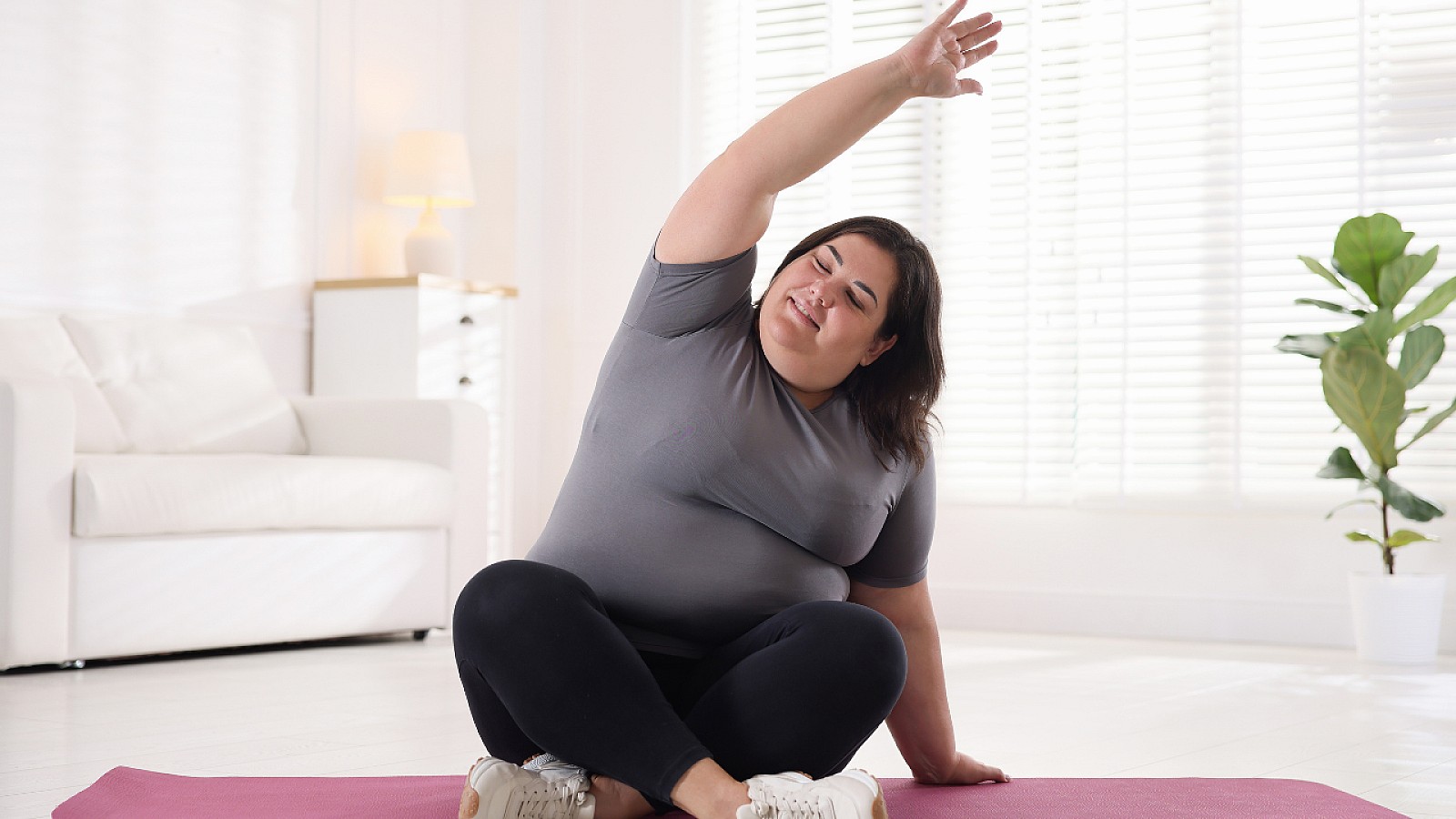 En fyldig kvinne sitter på yogamatte med en arm opp i sidebøy